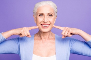 An older woman wearing a lavender cardigan points to her smile after seeing a dental implant dentist in Collierville